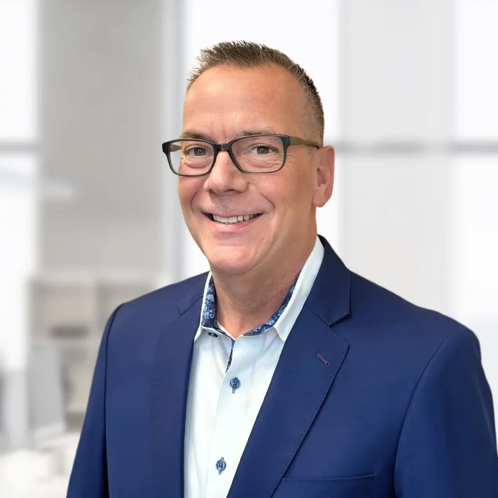 A man wearing glasses, a blue suit jacket, and a light-colored shirt smiles while standing in a bright office setting.