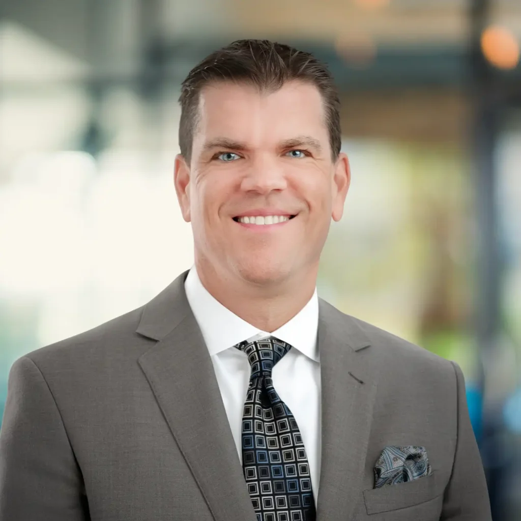 A man in a gray suit and patterned tie poses for a professional headshot against a blurred indoor background.