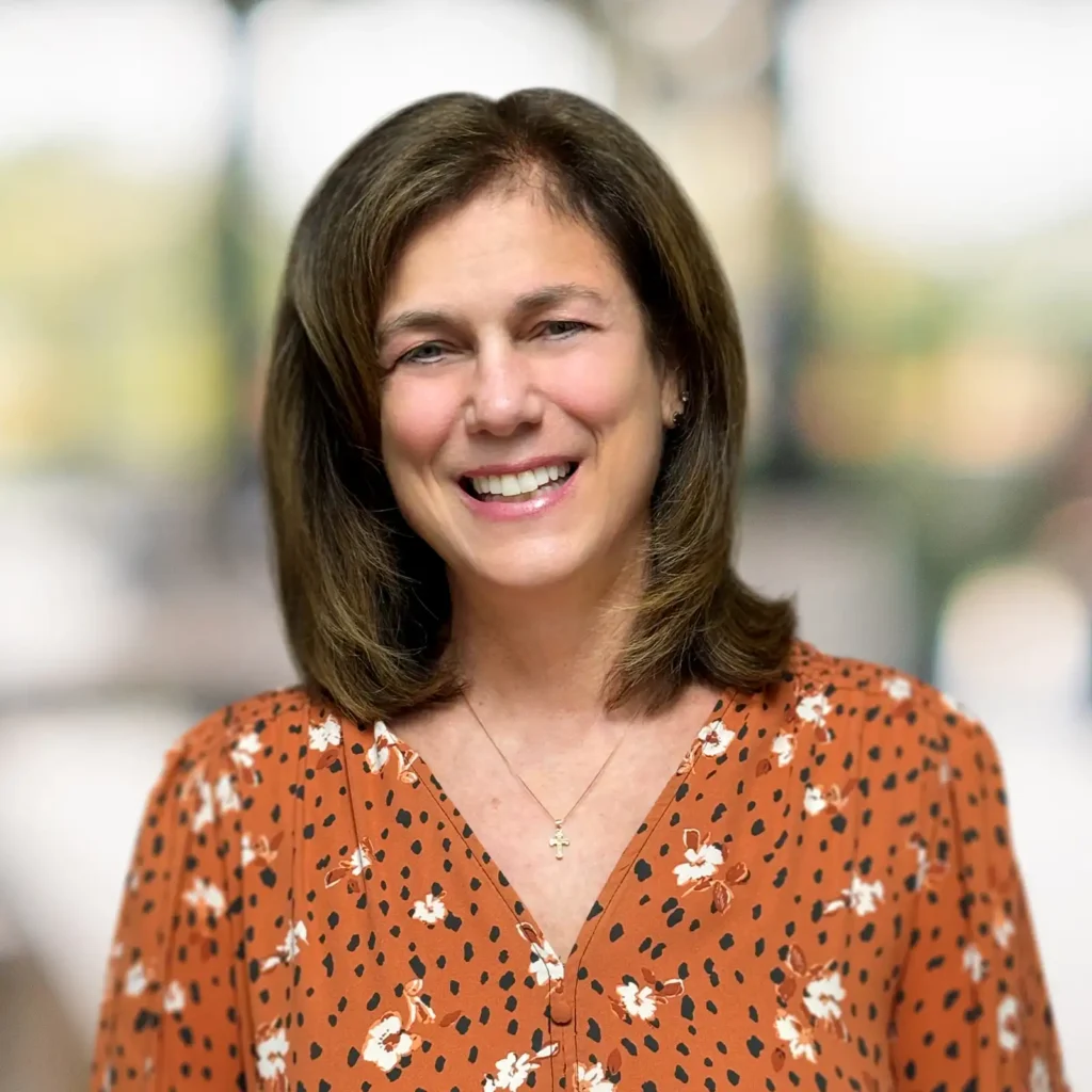 A woman with shoulder-length brown hair, wearing an orange floral blouse and a necklace, smiles at the camera with a blurred background.