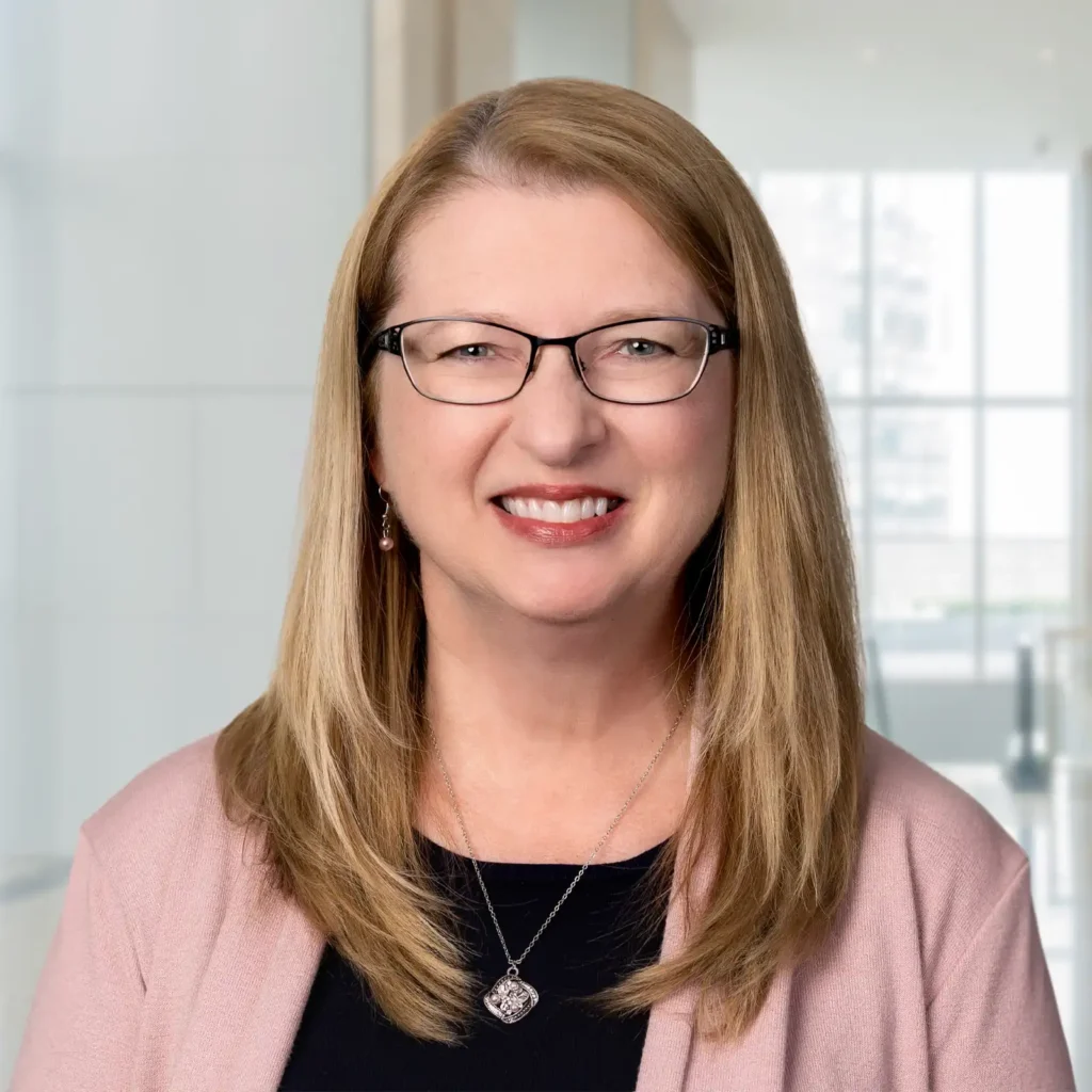 A woman with straight, blonde hair and glasses smiles at the camera. She is wearing a pink cardigan over a black top and a pendant necklace. The background is a bright, modern office.