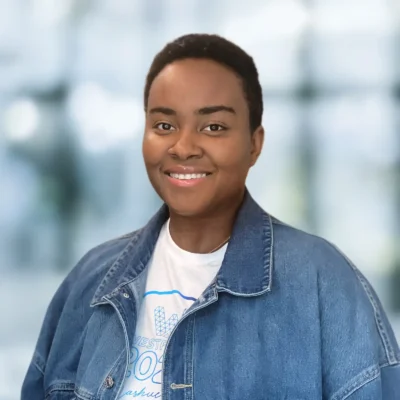 A person with short hair smiles at the camera, wearing a denim jacket over a white t-shirt, with a blurred background.