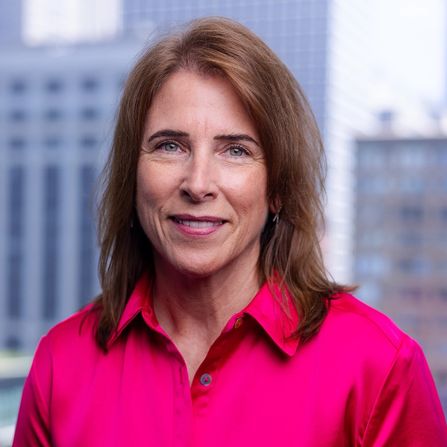 A woman with light brown hair and a pink blouse stands in front of a blurred cityscape background, smiling slightly at the camera.