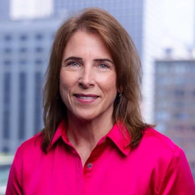 A woman with light brown hair wearing a bright pink button-up shirt smiles at the camera, with blurred office buildings in the background.