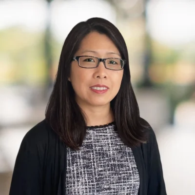 A woman with straight dark hair and glasses is wearing a black and white patterned top and a black cardigan, posing indoors with a blurred background.
