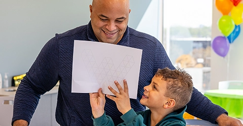 An adult and a child smile together as the child holds up a piece of paper with a geometric design. Colorful balloons are visible in the background.