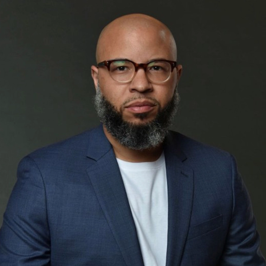 A man with a bald head, full beard, and glasses is wearing a blue suit jacket over a white shirt, posing against a dark background.