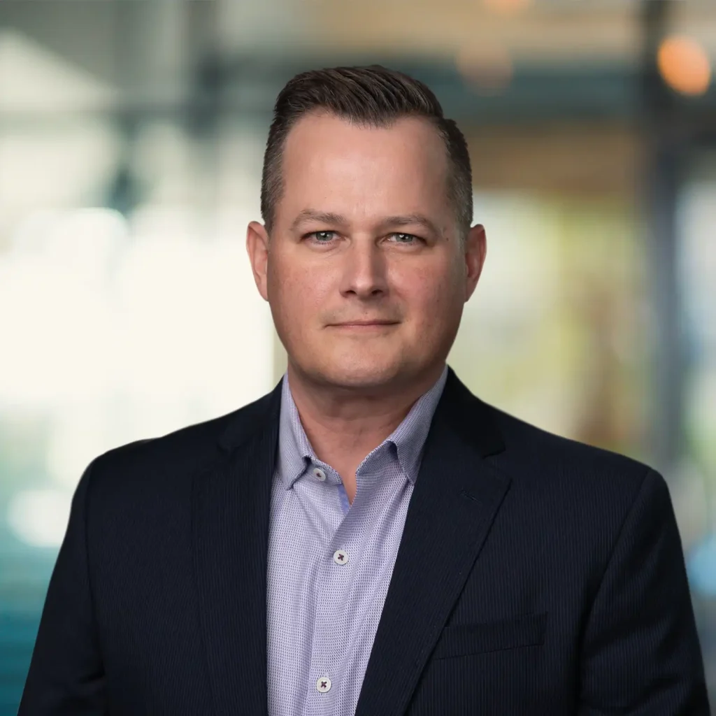 A man in a dark suit jacket and light shirt stands indoors, looking at the camera. The background is blurred with soft lighting and glass panels.