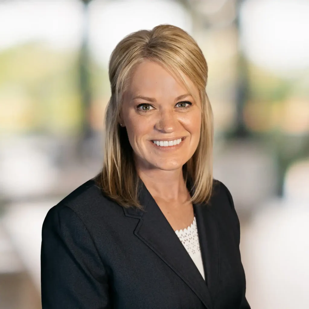 A woman with blonde hair in a dark blazer smiles at the camera with a blurred indoor background.