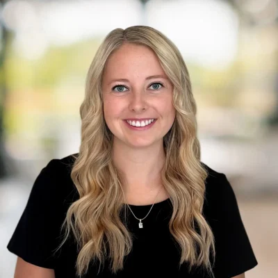A woman with long, wavy blonde hair and a black top smiles at the camera against a blurred background.