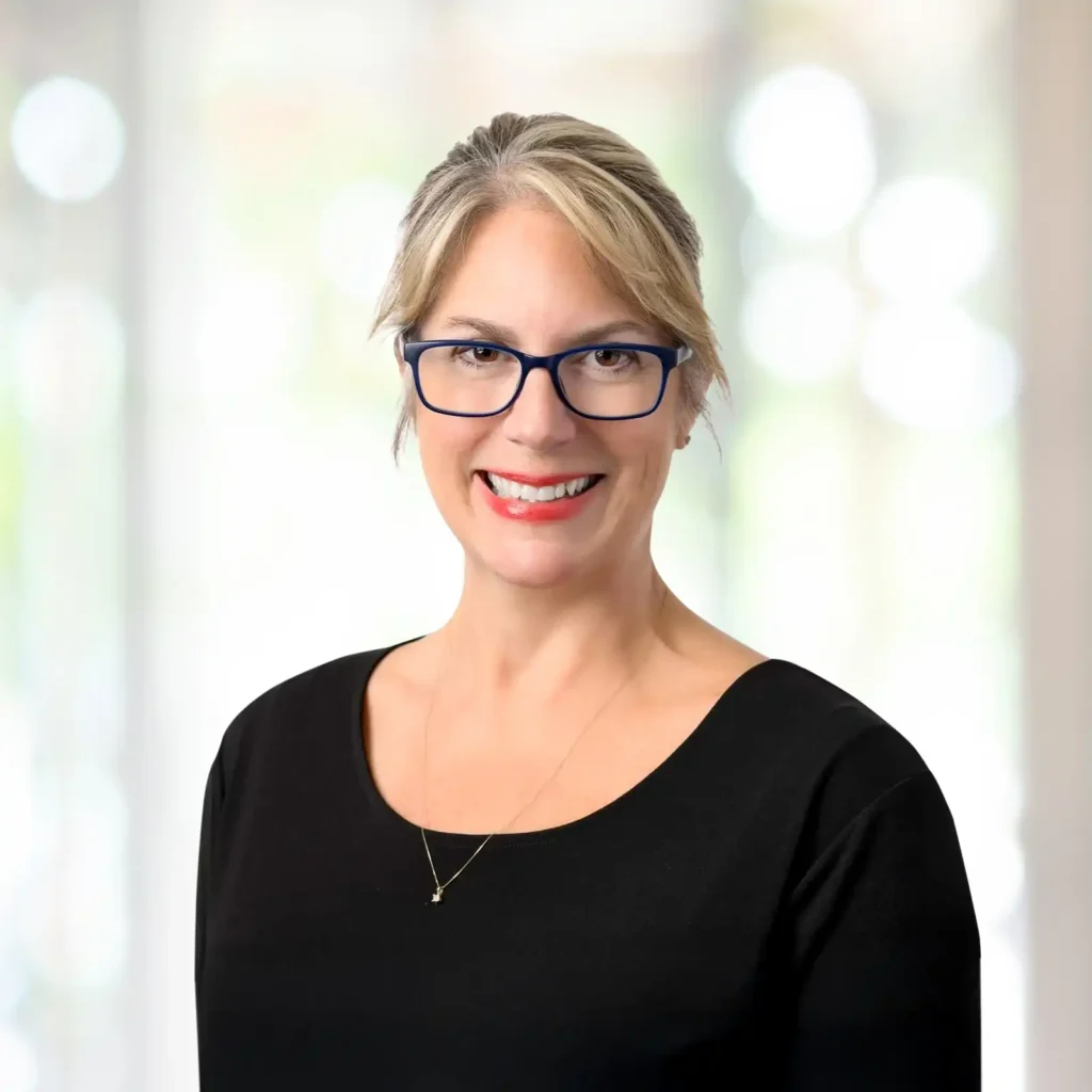 A woman with blonde hair, wearing glasses, a black top, and a necklace, smiles at the camera against a blurred light background.