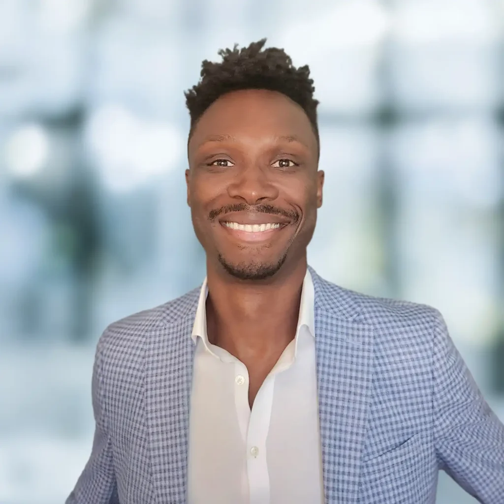 A man wearing a light blue checked blazer and white shirt smiles at the camera with a blurred background.