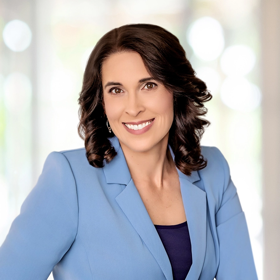 Woman with shoulder-length brown hair wearing a light blue blazer over a dark top, smiling, with a blurred light background.