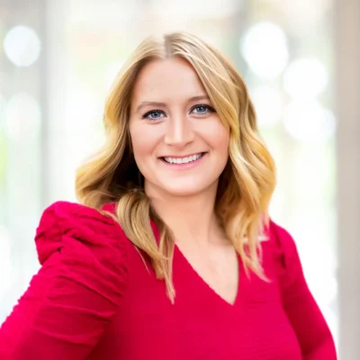 A woman with blonde hair wearing a bright red long-sleeve top smiles at the camera against a blurred, light-colored background.
