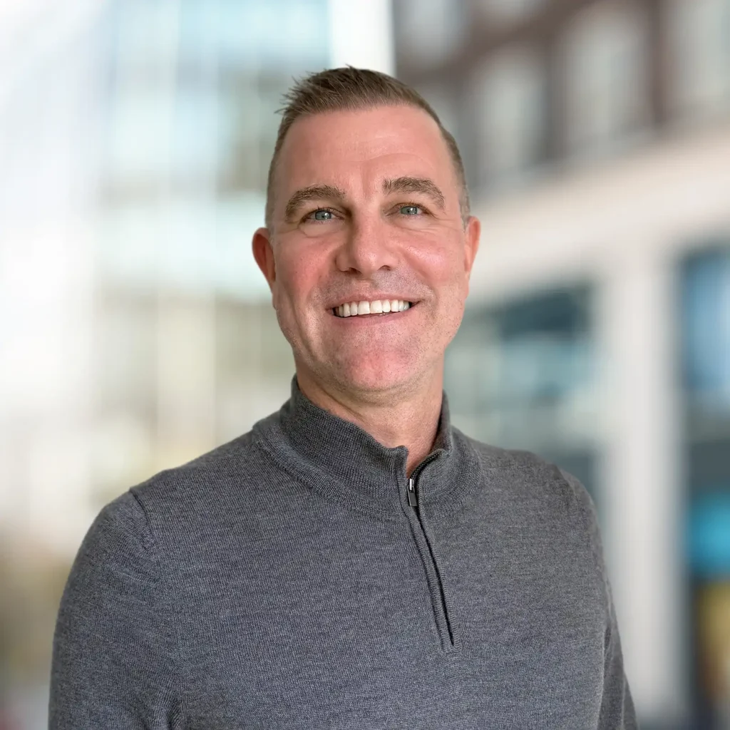 A man with short hair wearing a gray quarter-zip sweater stands smiling in front of a blurred urban background.