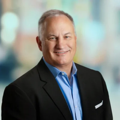 A middle-aged man in a dark suit jacket and light blue shirt smiles at the camera against a blurred background.