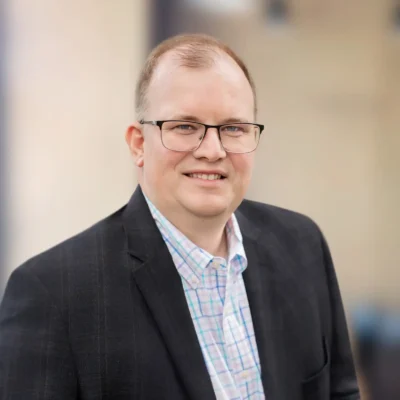 A man wearing glasses, a dark plaid blazer, and a checkered shirt stands and smiles in front of a blurred indoor background.