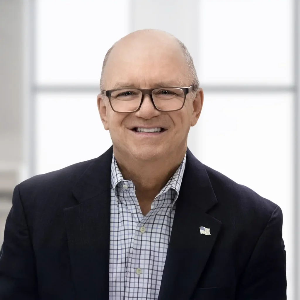 A smiling middle-aged man wearing glasses, a dark blazer, and a checkered shirt poses in front of a bright, blurred background.
