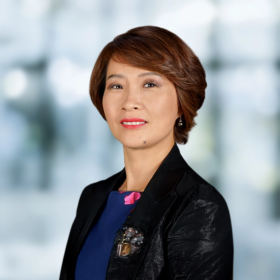 A woman with short brown hair wearing a dark blazer and blue top poses for a professional portrait against a blurred background.