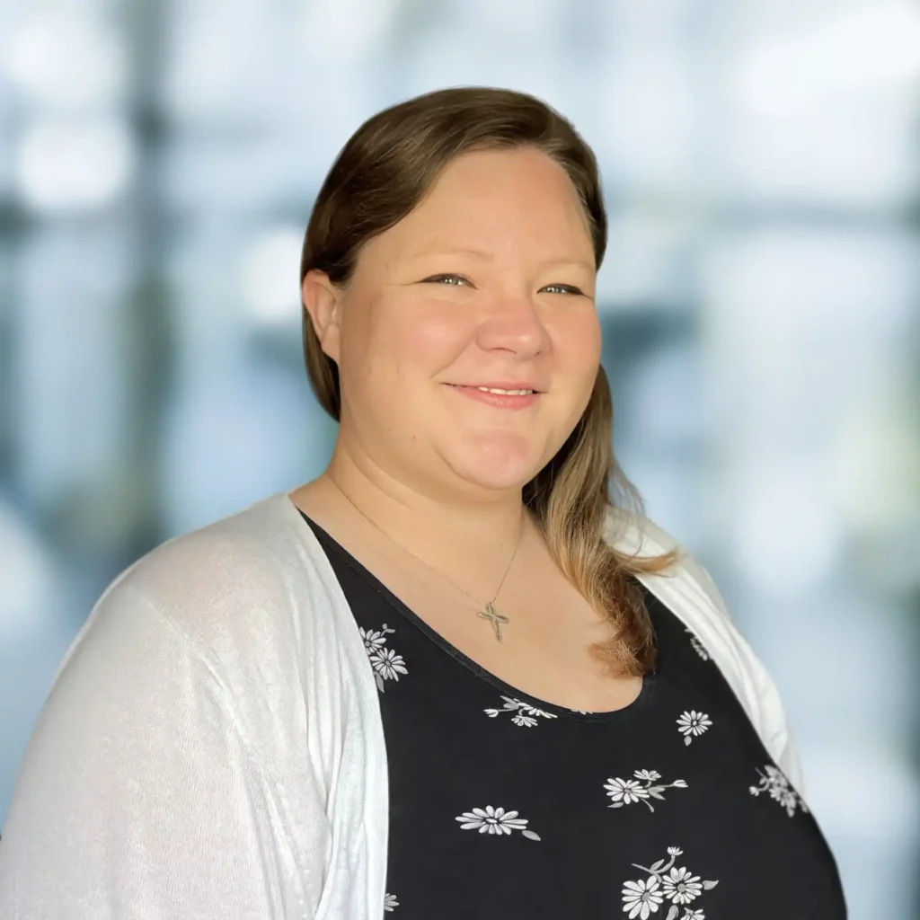 A woman with light brown hair wearing a black floral top, white cardigan, and a cross necklace, smiling in front of a blurred background.