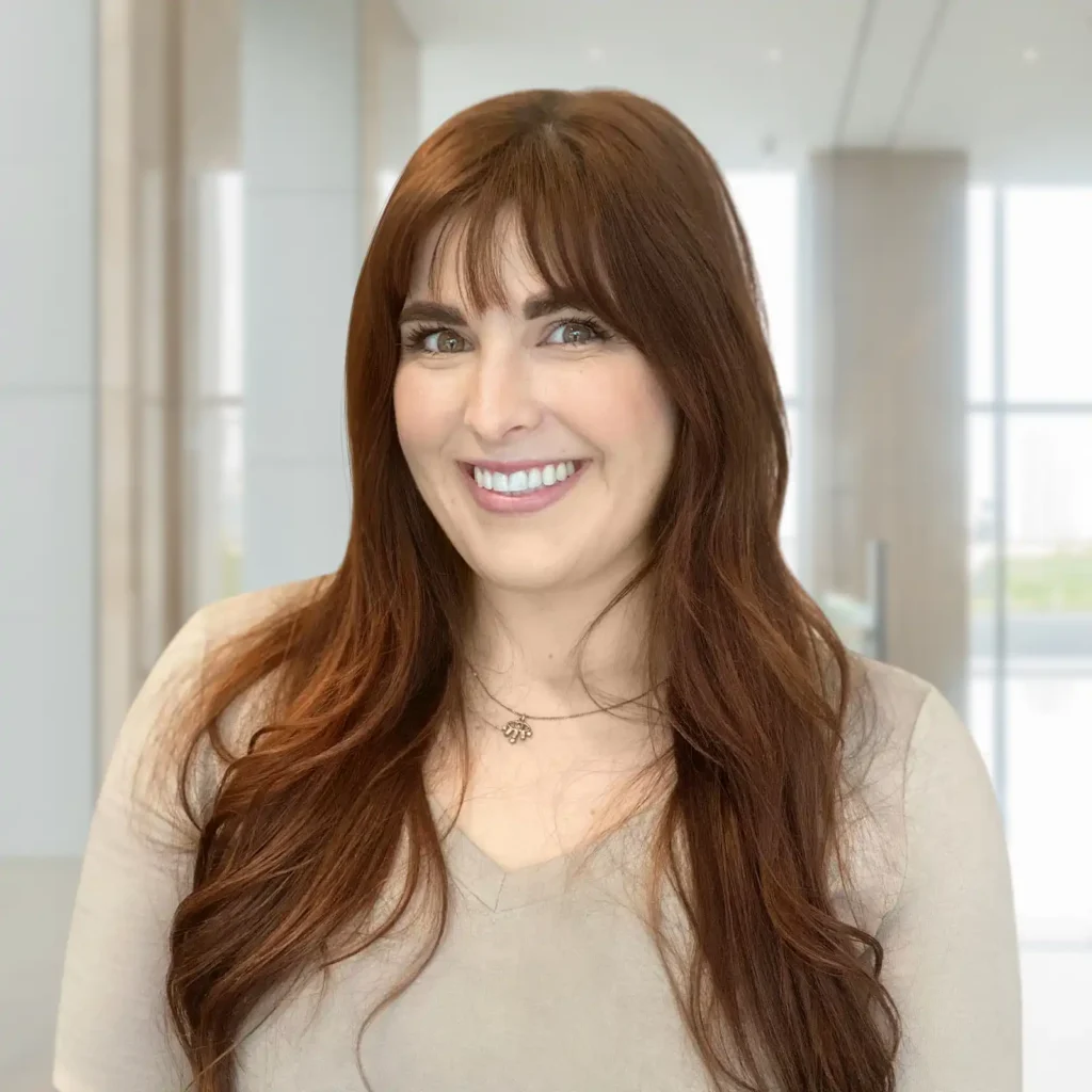 A woman with long brown hair and bangs, wearing a beige top, smiles while standing in a modern, bright office setting.