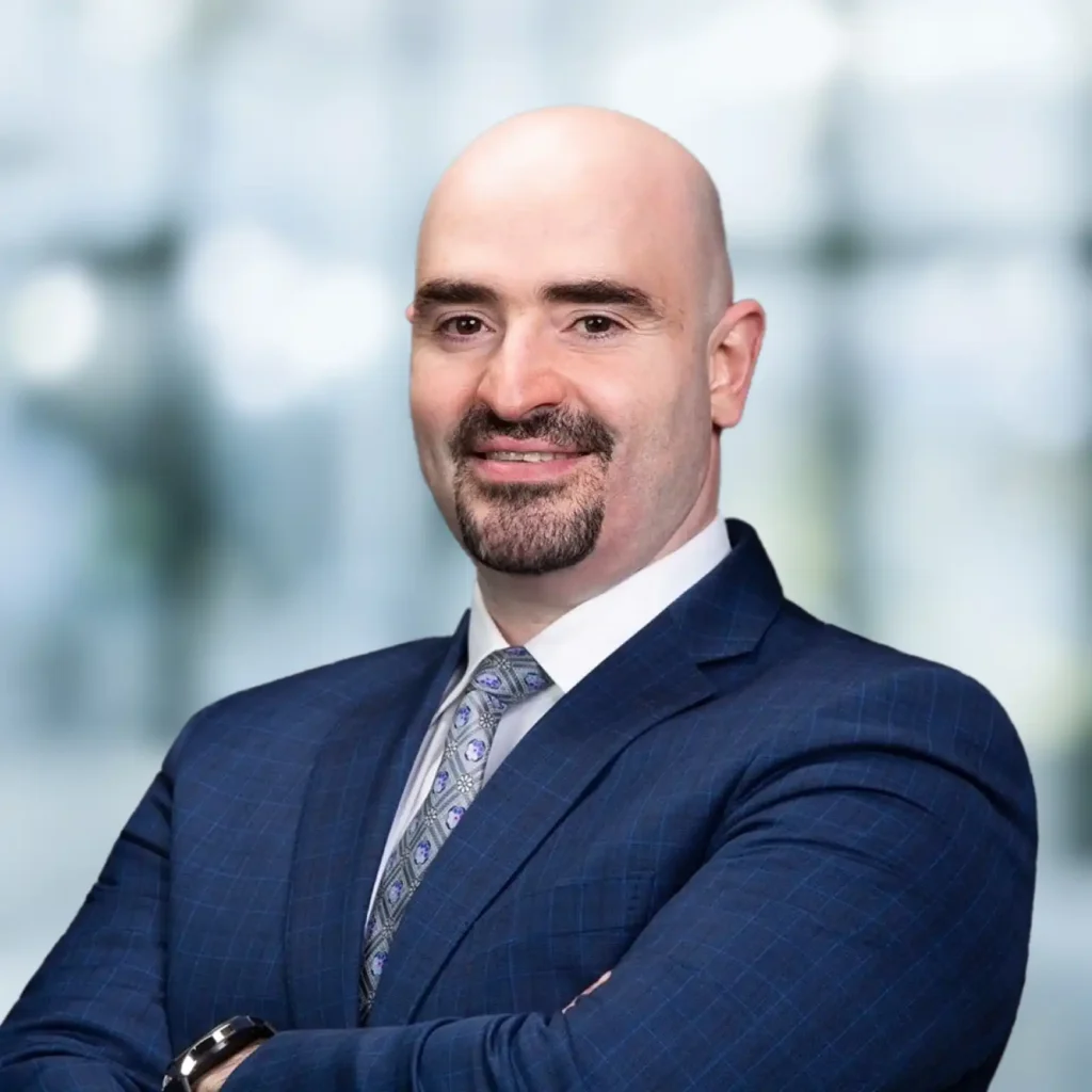A bald man with a goatee wearing a dark blue suit and patterned tie stands with arms crossed, smiling, against a blurred background.