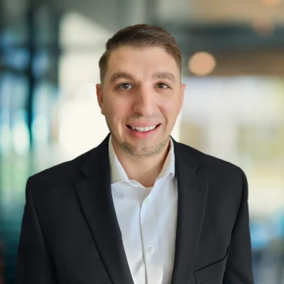 A man in a white shirt and black blazer stands indoors, smiling at the camera with a blurred background of windows and lights.