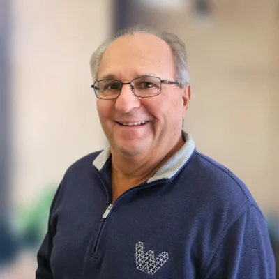 A middle-aged man with glasses and short gray hair is smiling, wearing a blue zip-up sweater with a geometric "W" logo, standing against a blurred background.
