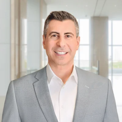 A man in a light gray suit and white shirt stands smiling in a modern, bright office setting with large windows.