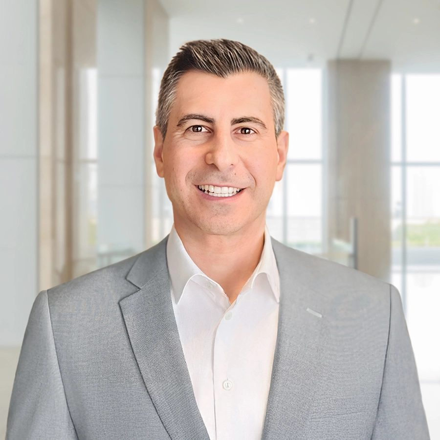 A man in a light gray suit and white shirt stands smiling in a modern, bright office setting with large windows.
