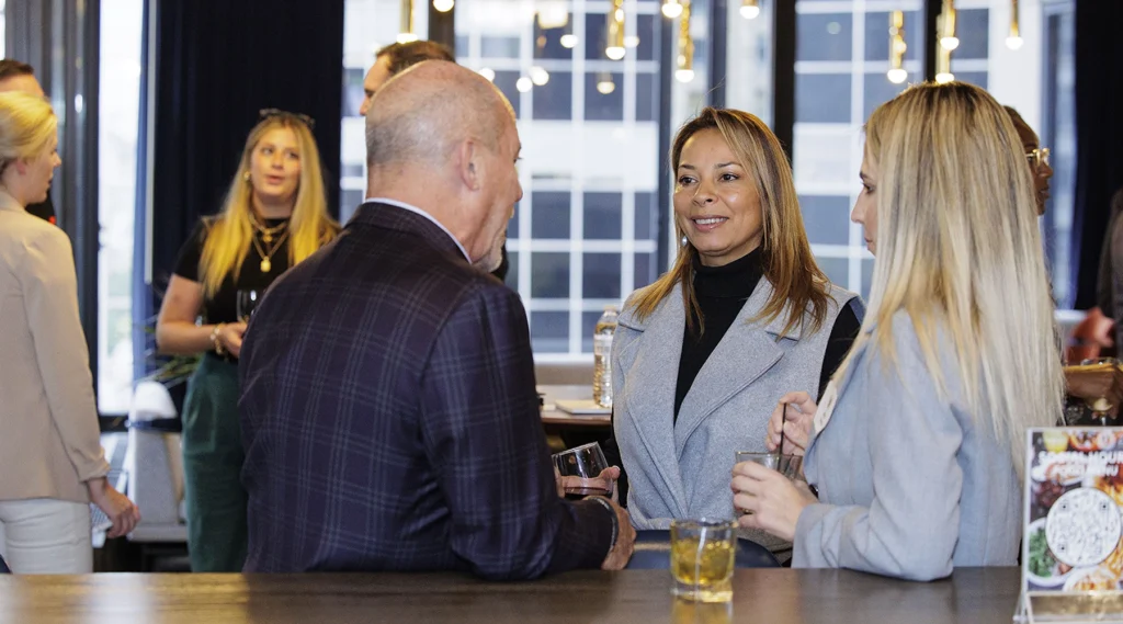 A group of people in business attire are conversing and holding drinks in a modern indoor setting with large windows.