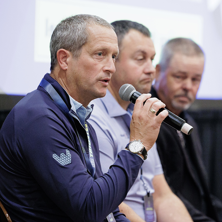 A man speaks into a microphone while seated on a panel with two other men, all wearing business casual attire.