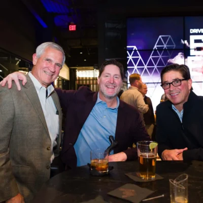 Three men in suits smile and pose together at a bar table with drinks, while other people socialize in the background at an indoor event.