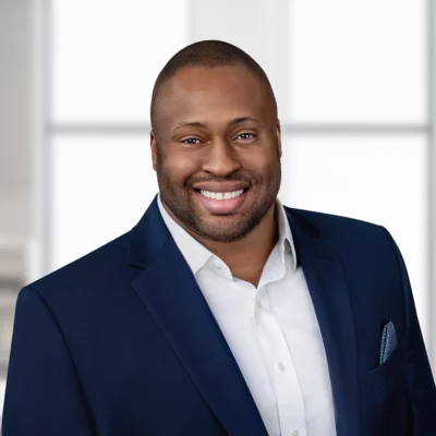 A man wearing a dark blue suit jacket and white shirt smiles at the camera in a bright, modern indoor setting.
