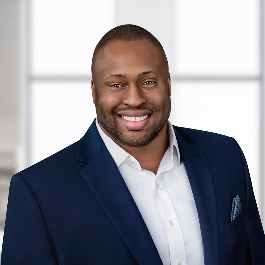 A man wearing a dark blue suit jacket and white shirt smiles at the camera in a bright, modern indoor setting.