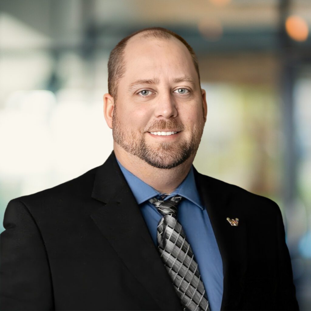 A man in a dark suit, blue shirt, and patterned tie stands indoors with a blurred office background.