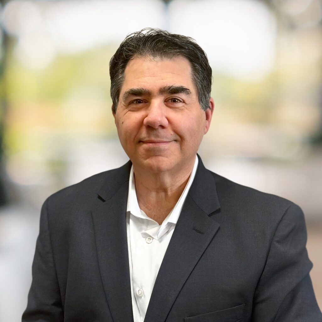 Middle-aged man with dark hair wearing a dark suit jacket and white shirt, standing in front of a blurred indoor background.