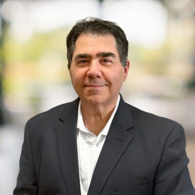 Middle-aged man with dark hair wearing a dark suit jacket and white shirt, standing in front of a blurred indoor background.