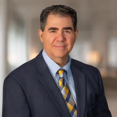 A middle-aged man in a dark suit, light blue shirt, and patterned tie poses in a blurred indoor setting.