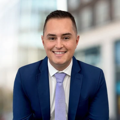 A man in a blue suit and light purple tie smiles at the camera with a blurred urban background.