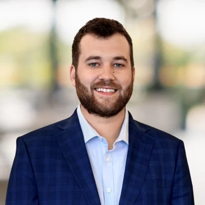 A man with a beard, wearing a blue suit jacket and light blue shirt, stands smiling in front of a blurred indoor background.