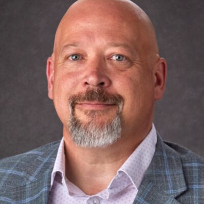 A bald man with a gray goatee wearing a plaid blazer and a light-colored shirt, posing against a plain dark background.