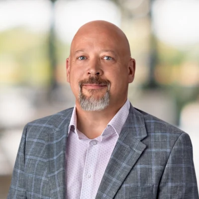 A bald man with a goatee and mustache, wearing a gray plaid blazer and light patterned shirt, stands against a blurred background.