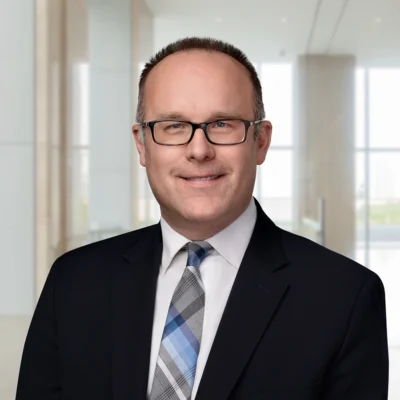 A man in a suit, tie, and glasses smiles at the camera while standing in a modern, brightly lit office space.