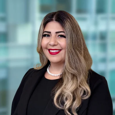 Woman with long, wavy blonde hair wearing a black blazer, black top, and pearl necklace, smiling at the camera against a blurred office background.