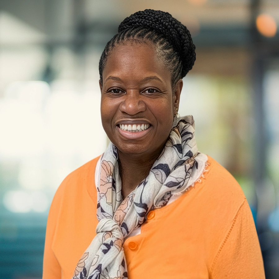 A woman with braided hair, wearing an orange sweater and a patterned scarf, smiles at the camera in a brightly lit indoor setting.