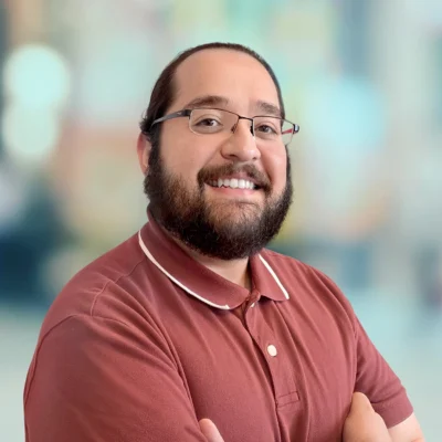 A man with glasses and a beard smiles, wearing a maroon polo shirt and standing with arms crossed against a blurred background.
