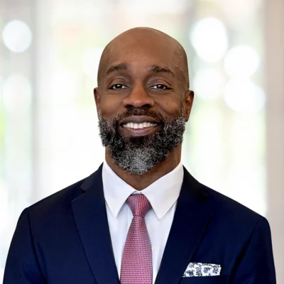 A man wearing a dark suit, white shirt, and pink patterned tie stands indoors, smiling at the camera. He has a short beard with some gray hair.