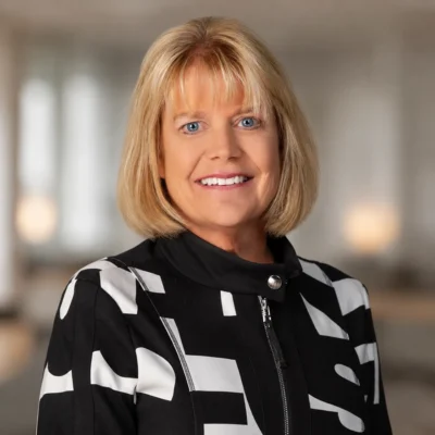 A woman with blonde hair wearing a black and white patterned jacket, smiling, stands in front of a softly blurred indoor background.