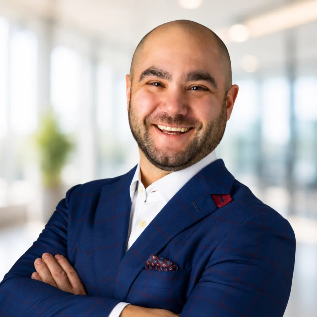 A smiling man in a blue suit stands with arms crossed in a bright, modern office setting.