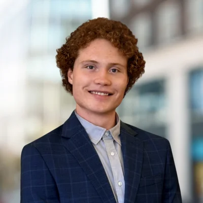 A young person with curly red hair, wearing a blue plaid suit jacket and light shirt, smiles at the camera with a blurred office background.